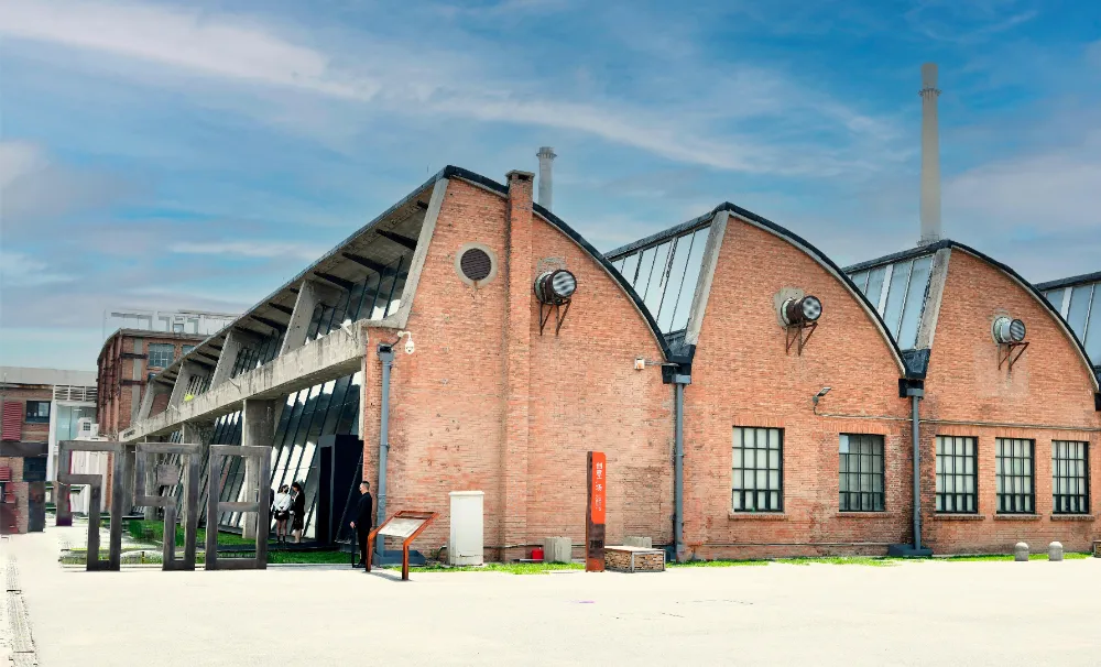 A heavy industrial steam pipe painted matte grey running across a red-brick Bauhaus factory building facade inside the 798 Art Zone.