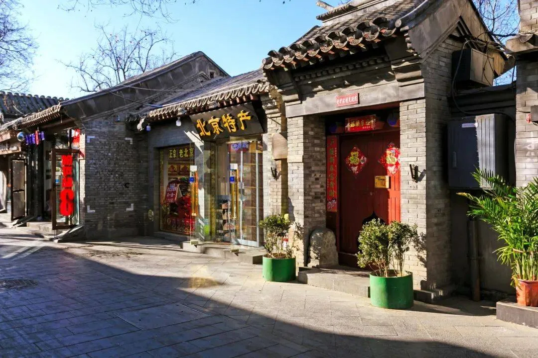 An elderly resident sitting on a wooden chair outside a traditional grey brick courtyard gate in a Beijing hutong.