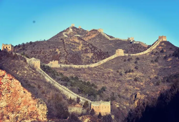A crumbling watchtower on the unrestored Jiankou Great Wall, with wild vegetation growing between weathered Ming dynasty bricks under a soft sky.